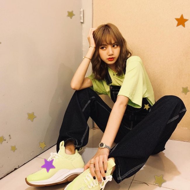 Looks da Lisa do BLACKPINK Lisa, integrante do grupo Blackpink, sentada com uma das mãos tocando seu pé e a outra apoiada na cabeça; com uma expressão pensativa;usando camiseta verde clara, assim como seu tênis e calça jeans.