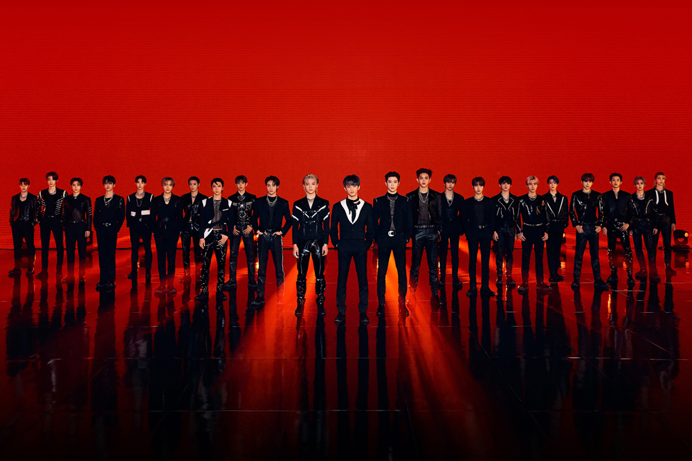 NCT_Divulgacao Supergrupo NCT em um fundo vermelho, os integrantes estão alinhados na diagonal formando uma espécie de triângulo com um integrante no meio; todos vestem roupas completamente pretas, algumas com detalhes brancos