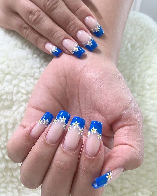 Duas mãos exibindo as unhas com base clara e francesinha azul com florzinha branca.