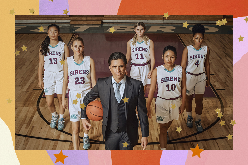 Elenco de BIG SHOT, vestido no uniforme de basquete da série, posicionadas em forma de triângulo em quadra, com o treinador interpretado por John Stamos na pontoa segurando uma bola de basquete.