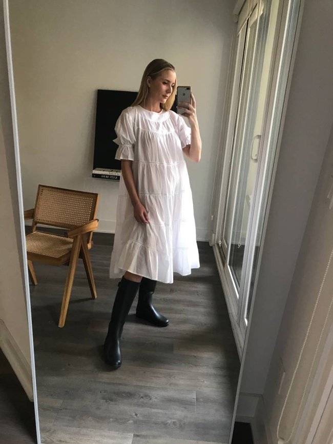 botas-do-inverno-2021-galocha Jovem posando em frente a espelho com vestido e bota preta. Podemos ver uma televisão e parte de uma cadeira atrás dela, que posa para foto no espelho.