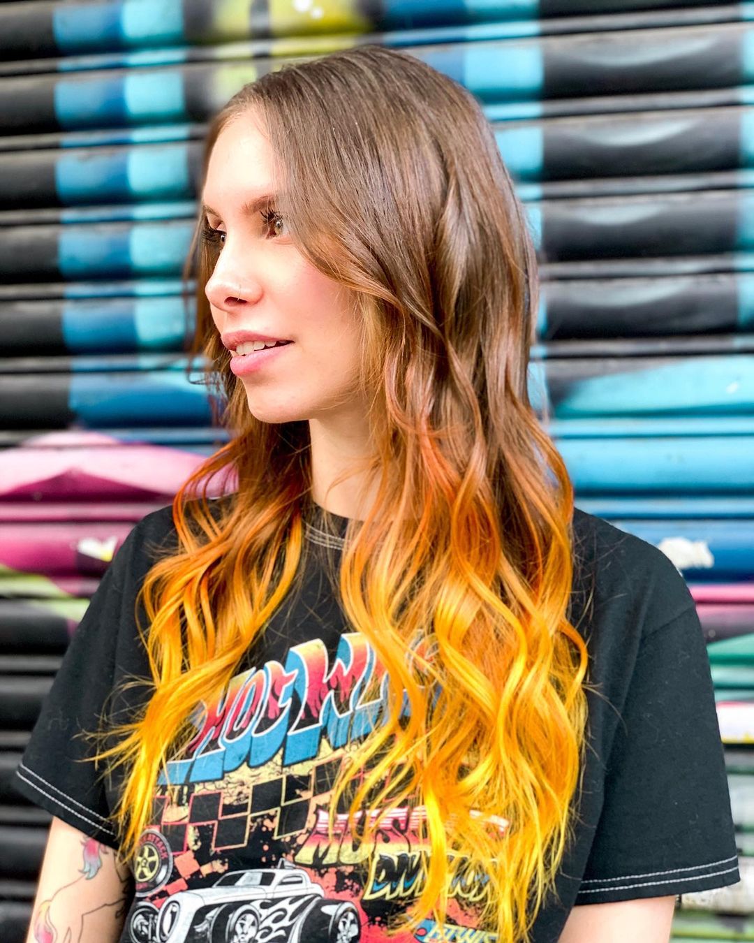 cabelo-colorido-amarelo Mulher de perfil olhando para a direita. Ela está vestindo uma camiseta preta com uma estampa de letras coloridas, cabelo solto, ondulado e com as pontas amarelas