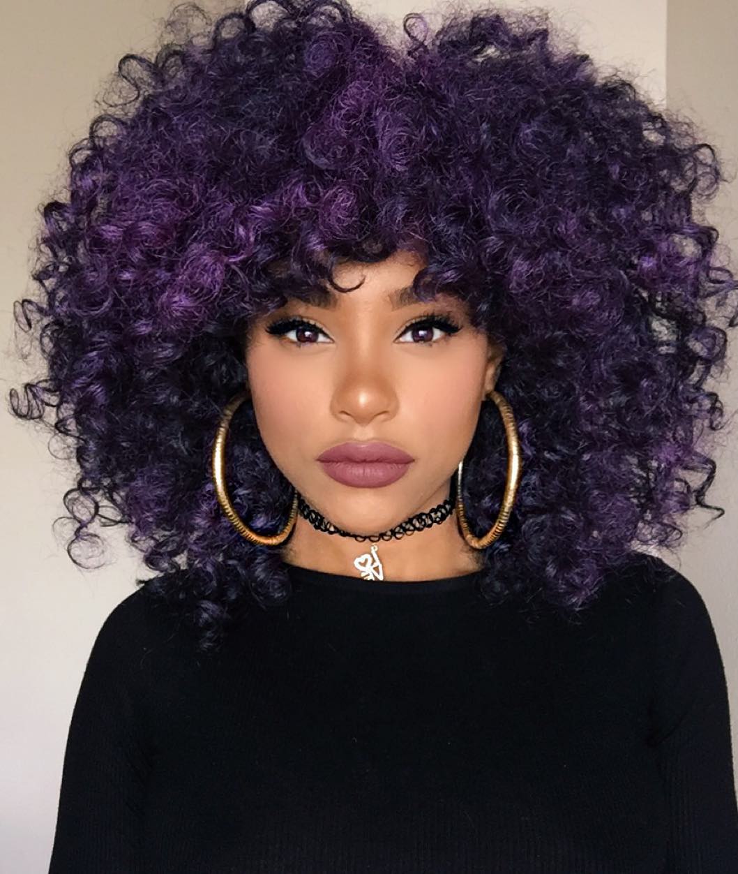 Foto de uma mulher. Ela está vestindo uma blusa de manga longa preta, brinco de argola dourado, chocker preta e batom marrom. O cabelo dela é roxo escuro, ela olha para a câmera mas não sorri.