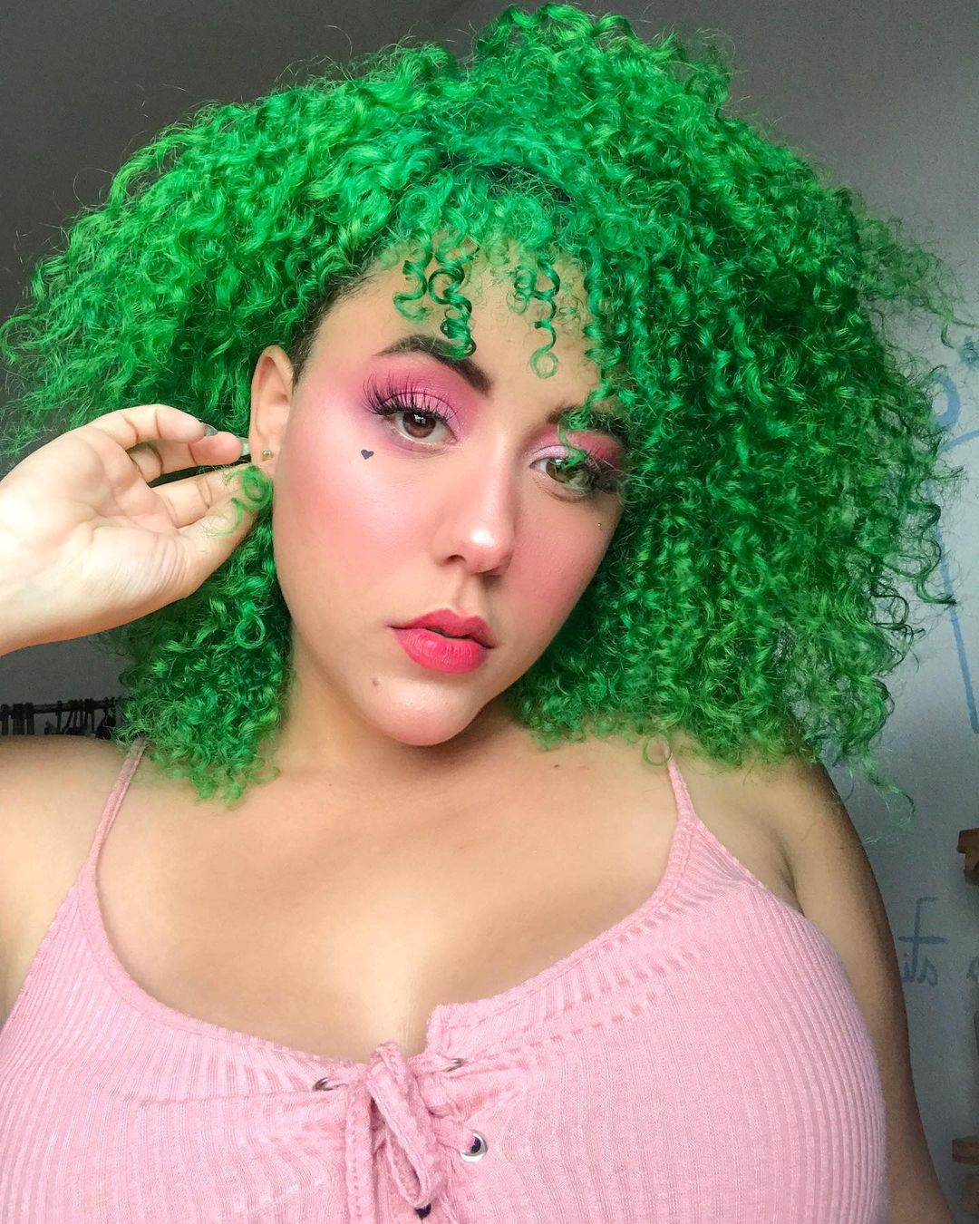 cabelo-colorido-verde-cacheado Selfie de uma mulher. Ela está vestindo uma blusa de alcinha na cor rosa claro, está com o batom e maquiagem rosa, cabelo é cacheado, totalmente verde e está solto. A mão direita da mulher está segurando o cabelo atrás da orelho e ela olha para a câmera sem sorrir.