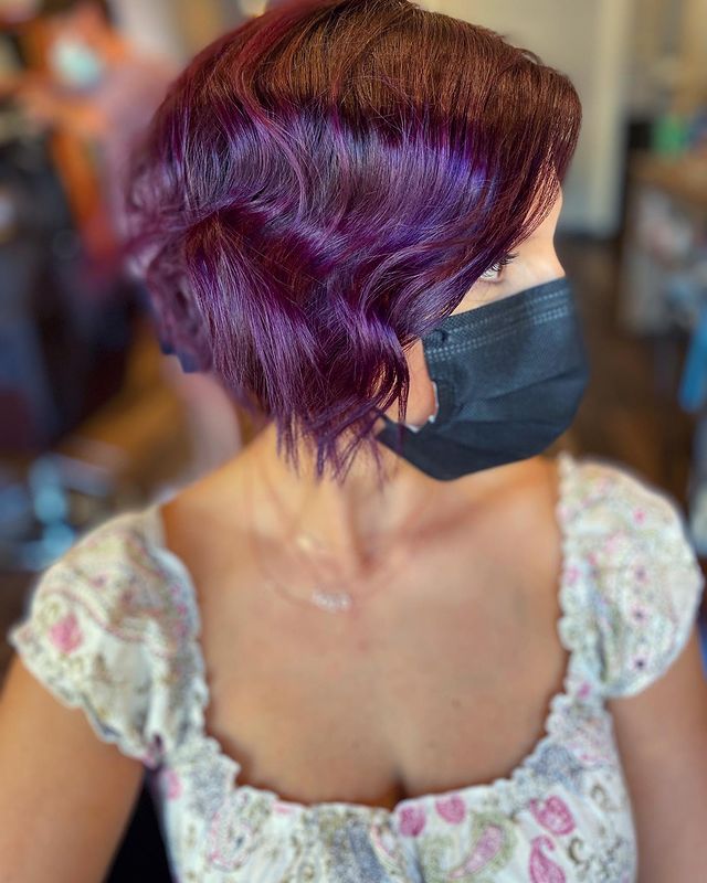 Jovem olhando para o lado co cabelo curto e roxo, usando blusa de babados branca com flores, não conseguimos ver sua expressão pois ela usa uma mascara de proteção preta.