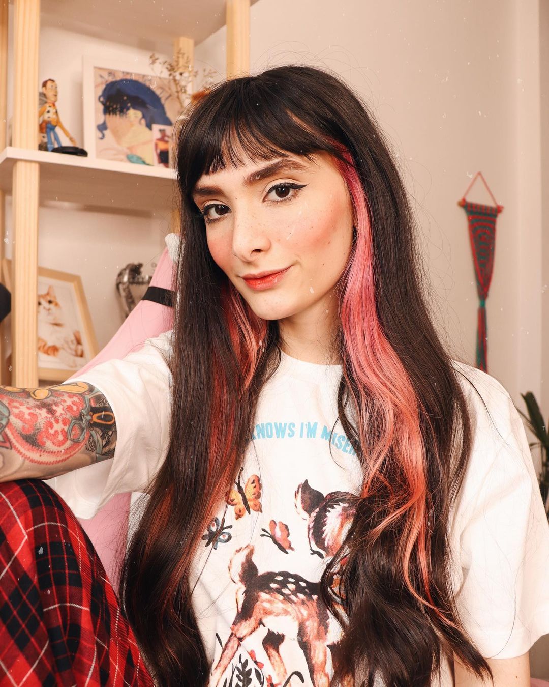 cabelo-mecha-lateral-rosa Selfie de uma mulher. Ela está vestindo uma camiseta branca com estampa de borboleta, calça com estampa xadrez preta e vermelha, cabelo preto com franja solto e com mechas laterais na cor rosa pastel. A mulher sorri para a câmera.
