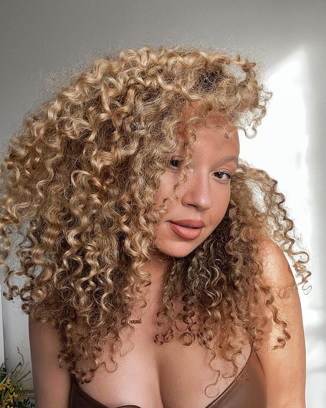 cabelo_loiro_cacheado Selfie de uma mulher. Ela usa um top marrom e cabelo cacheado solto e loiro. Ela olha para a câmera e não sorri.