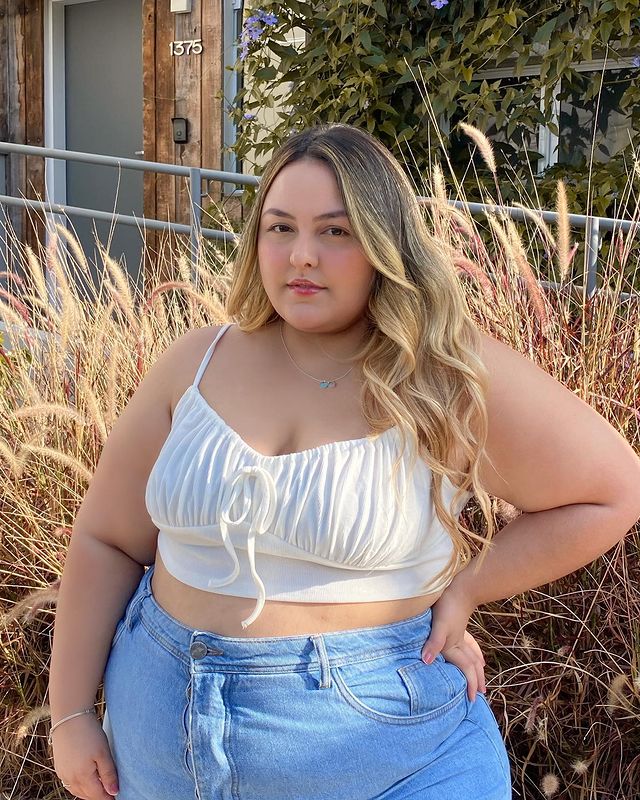 cabelo_loiro_champanhe Foto de uma mulher. Ela usa um top cropped branco e calça jeans. Ela está de cabelo solto, mão esquerda na cintura, olha para a câmera e não sorri.