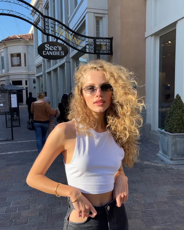 cabelo_loiro_ondulado Foto de uma mulher na rua. Ela usa um top cropped branco, calça preta e está com o cabelo ondulado solto. Ela olha para a câmera e não sorri.