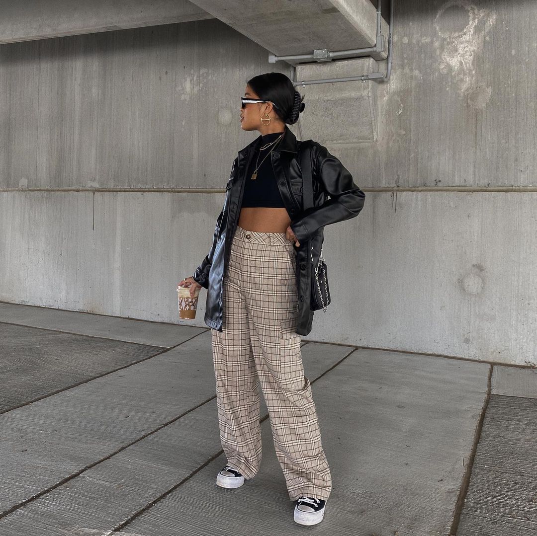 calca_xadrez Foto de uma mulher em um estacionamento. Ela usa um cropped preto, jaqueta de couro preta, calça xadrez branca com marrom, tênis preto e bolsa preta. Ela olha para o lado e segura um copo de café.