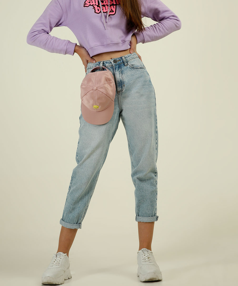 colecao-capricho-marisa-04 Garota usando calça mom jeans da coleção da CAPRICHO com a MARISA. Tem um boné rosa pendurado em um dos passadores da calça, ela está de tênis branco e dá para ver um pedaço do moletom lilás. Suas duas mãos estão na cintura.