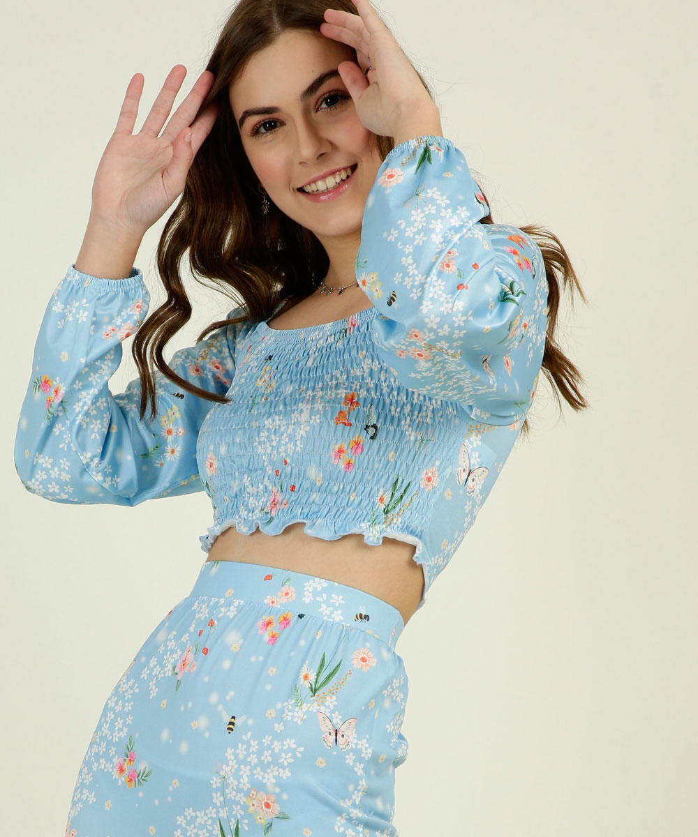 Garota usando blusa cropped de manga longa com estampa floral e fundo azul da coleção da CAPRICHO com a MARISA. Ela também está usando a saia, que só dá para ver uma parte, na mesma estampa. Ela está com as duas mãos no cabelo e sorrindo.