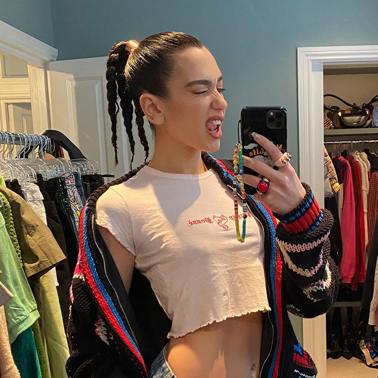 cordinha-celular-dua-lipa Dua Lipa usando top cropped branco e cardigan colorido em frente ao espelho. Ela está de lado, segurando o celular que tem uma cordinha de miçangas pendurada, e fazendo uma careta piscando o olho e mostrando a língua.
