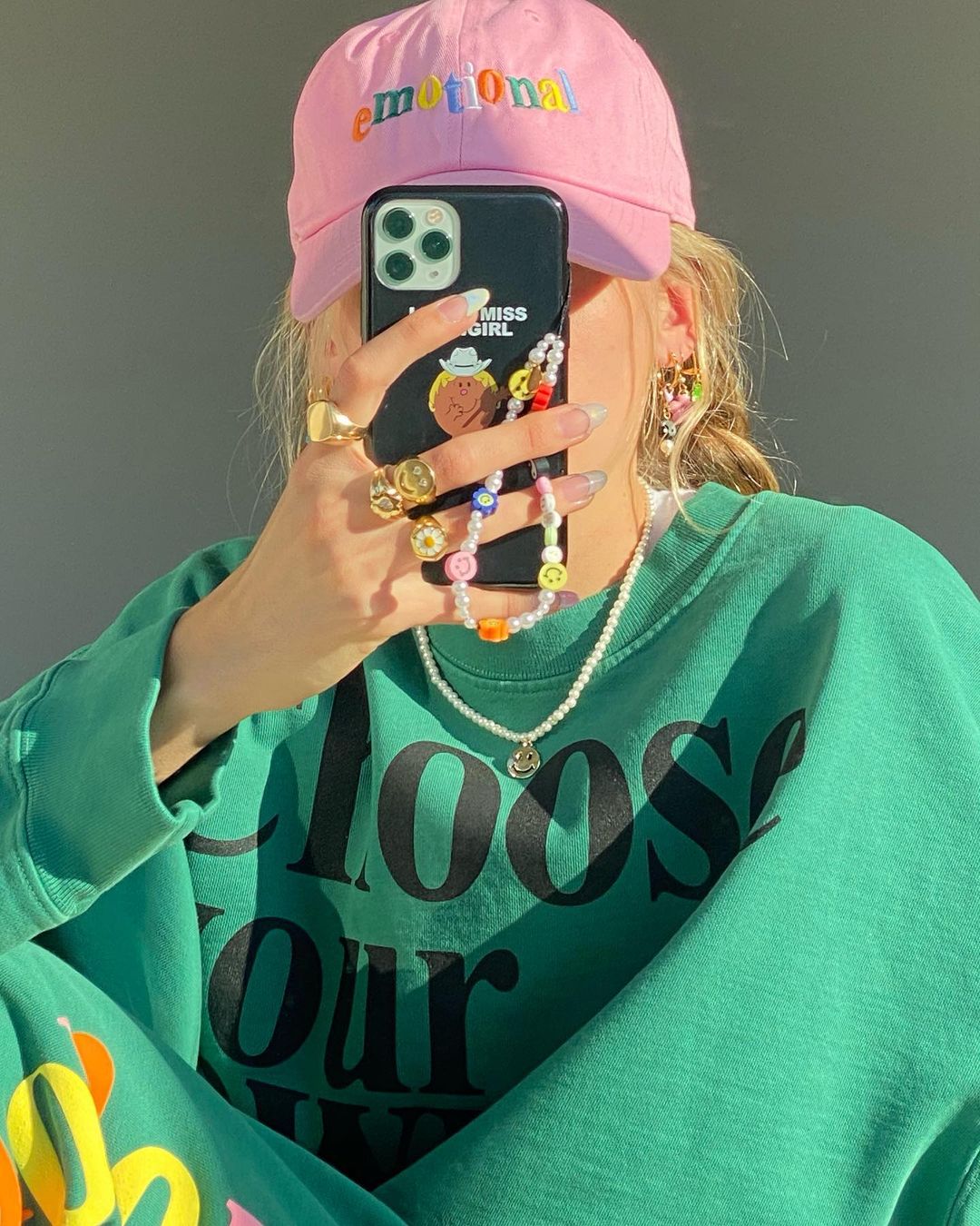 cordinha-celular-laurenladnier Garota usando boné rosa, moletom verde enquanto tira foto em frente ao espelho. Ela está cobrindo o rosto com seu celular, que tem uma cordinha de miçangas pendurada.