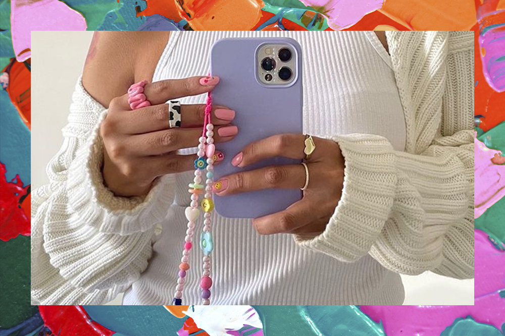 Garota usando regata branca, cardigan off white e tirando foto na frente do espelho segurando o celular que tem uma cordinha de miçangas pendurada. No close da foto, só dá para ver a região da barriga e do tórax. Ao fundo da montagem, há um efeito de várias tintas coloridas.