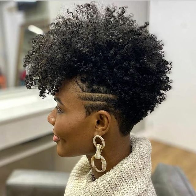curto crespo com a lateral raspada Jovem sentada de lado, não conseguimos ver seu rosto, usando brincos longos, com blusa de gola rolê branca, sentada em uma cadeira cinza, exibindo a lateral da cabeça raspada.