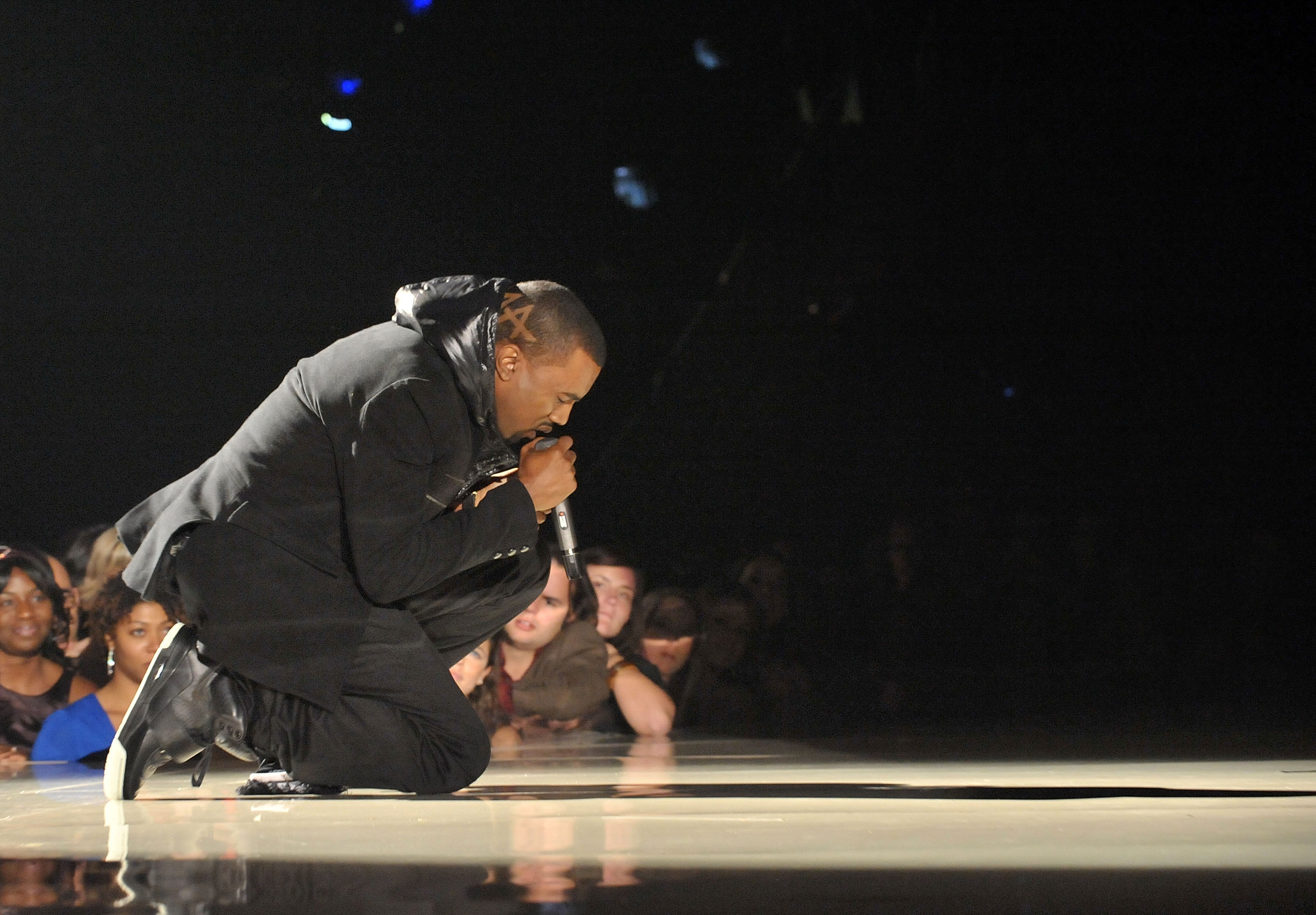 Kanye West no palco do Grammy de 2008. Ele veste um casaco com capuz preto, calça preta e tênis de couro preto com cano alto. Ele está ajoelhado e cantando com o microfone na mão direita.