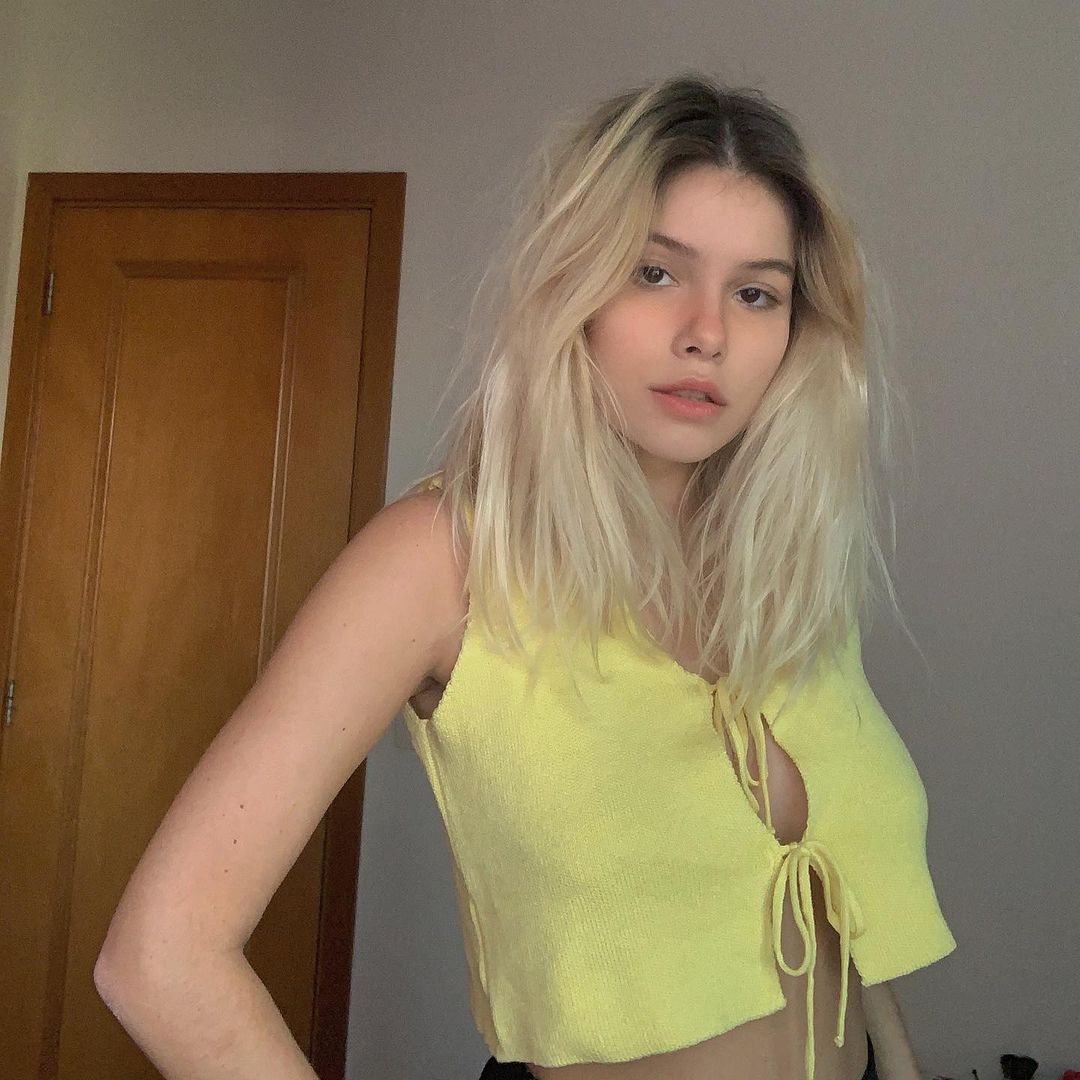 loiro_perolado Foto de uma mulher. Ela usa um top amarelo e cabelo solto. Ela olha para a câmera, não sorri e está com a mão direita apoiada na cintura.