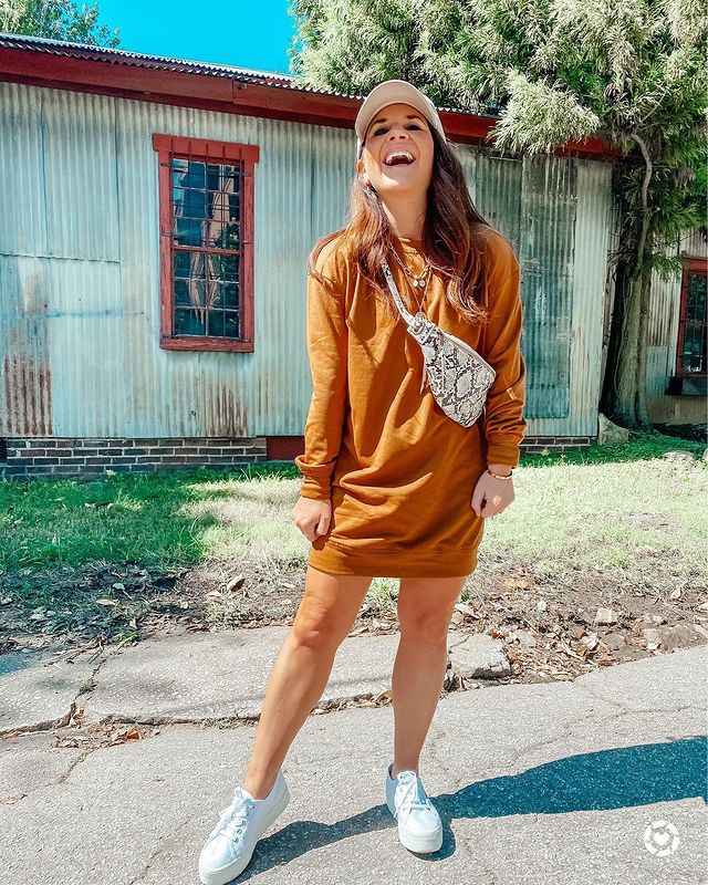 look-blusa-moletom-vestido-addictedto2dayshipping Garota usando blusa de moletom mostarda como vestido, tênis branco e pochete com estampa de cobra atravessada no corpo, além de um boné bege na cabeça. Ela está dando risada e olhando para cima.