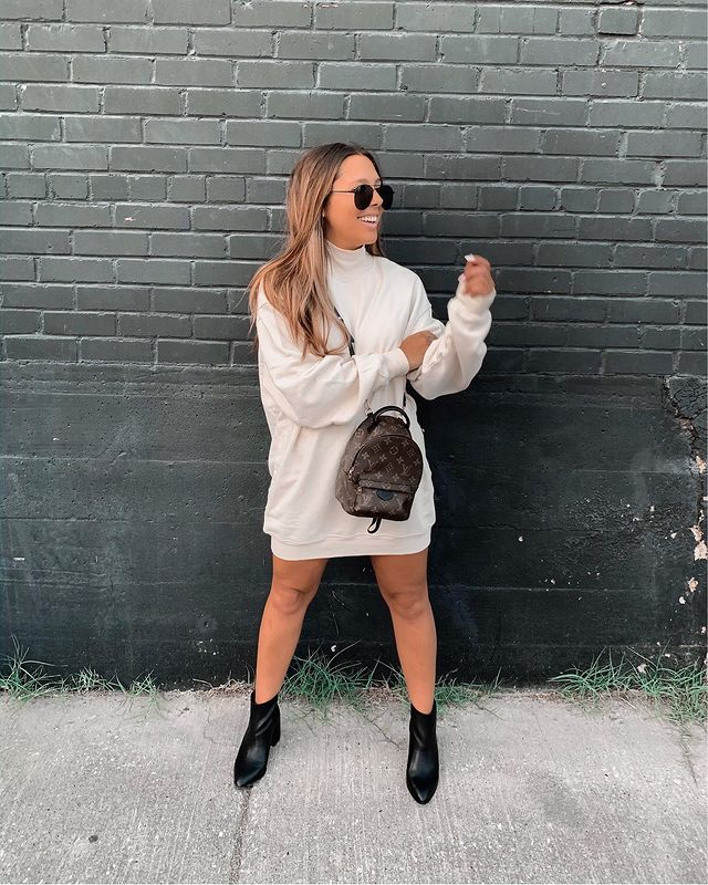 look-blusa-moletom-vestido-ashleylarea Garota usando blusa de moletom bege como vestido, bota preta de cano curto com bico fino, minimochila Louis Vuitton atravessada no corpo e óculos de sol. Ela está olhando para o lado, sorrindo, com uma das mãos apoiada no outro braço.