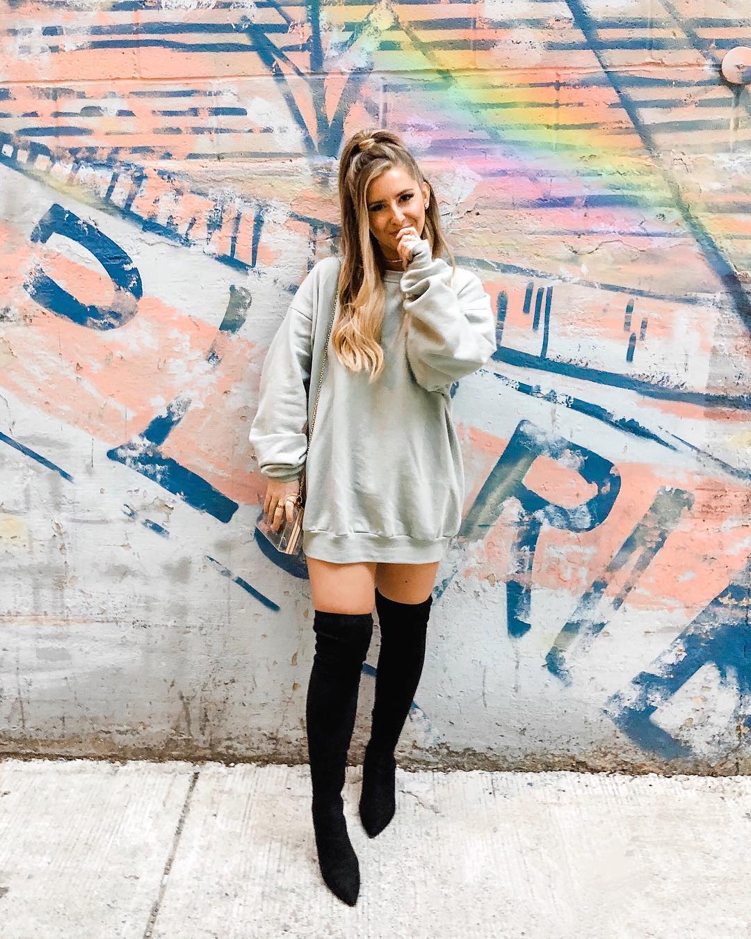 look-blusa-moletom-vestido-chloandtell Garota usando look com vestido de moletom verde claro, bota over the knee preta, bolsa pendurada em um dos ombros e penteado semipreso. Ela está com uma das mãos cobrindo a boca.