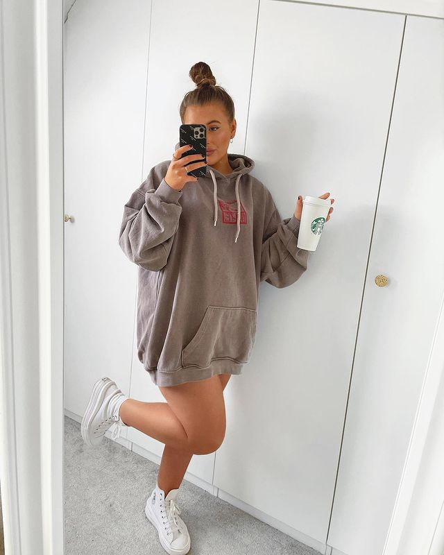 look-blusa-moletom-vestido-looksbylol Garota em frente a um espelho tirando foto com o celular em uma das mãos e um copo branco de café na outra. Ela está usando uma blusa de moletom marrom como vestido e um tênis branco de cano alto, e uma das pernas está dobrada para trás.