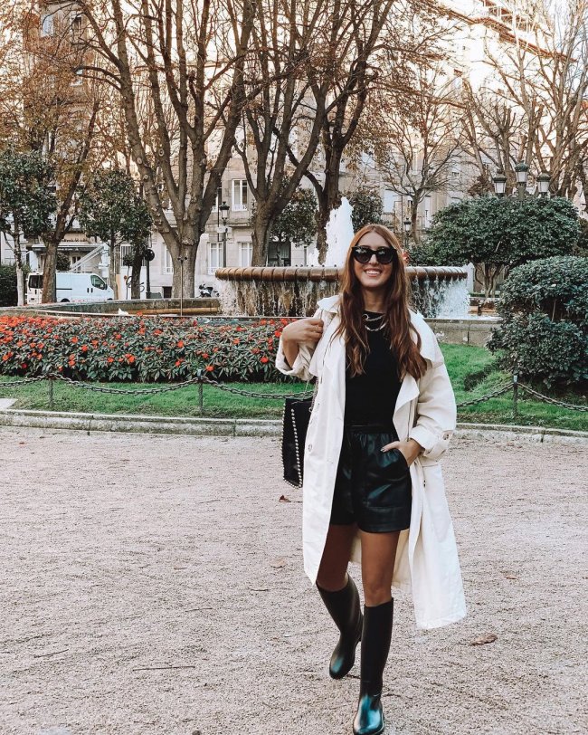 look com galocha Jovem com sobretudo branco, meia calça, bota preta, short e blusa, usando óculos preto, com uma mão no bolso.