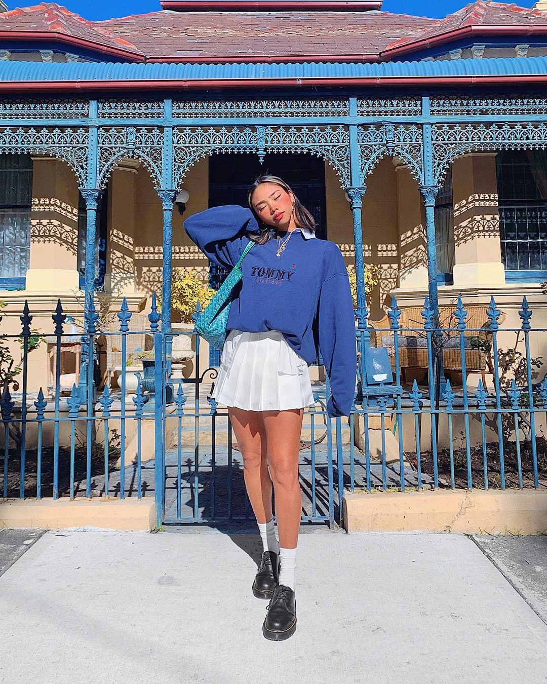 look_moletom_vintage_saia_colegial Foto de uma mulher em frente a uma casa com o portão azul. Ela usa um moletom vintage azul, saia colegial branca, sapato preto com meia branca. Ela está de olho fechado e cabeça apoiada no braço direito.