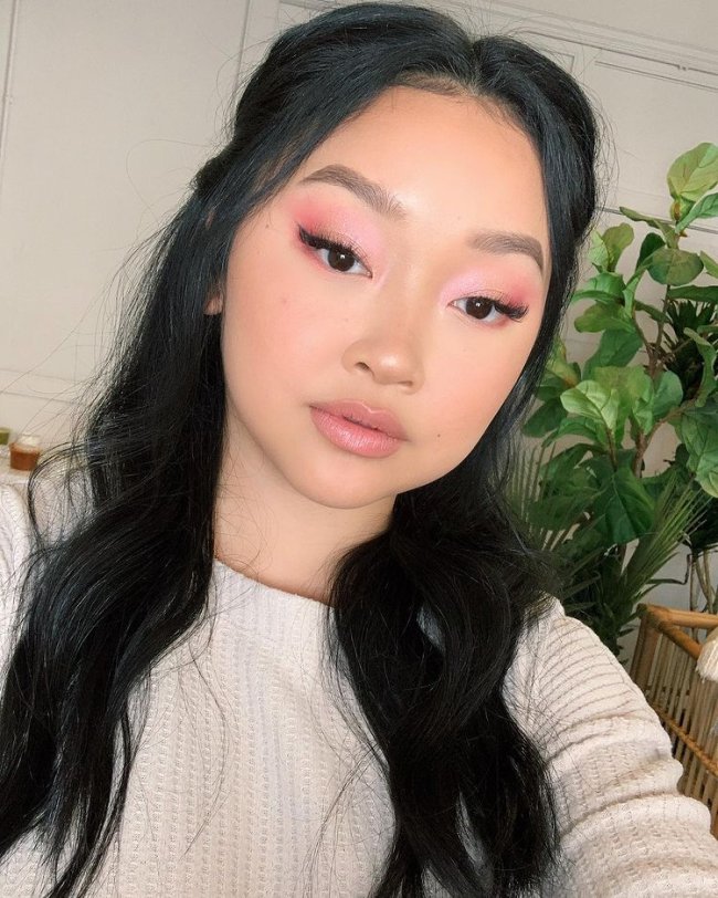 Selfie da atriz Lana Condor. Ela usa uma blusa branca, cabelo meio preso e maquiagem com sombra rosinha e delineado gatinho. Ela olha para a câmera e não sorri.