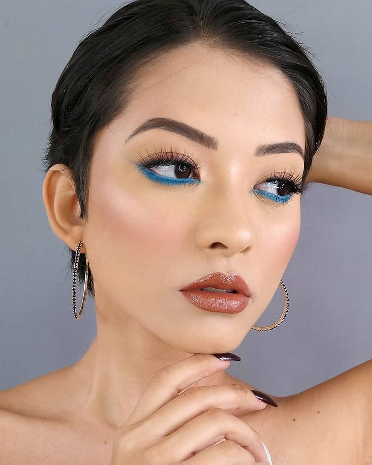 maquiagem-delineado-invertido-raissalisboabeauty Garota usando maquiagem com delineado azul invertido rente aos cílios inferiores. Ela tem o cabelo curtinho, está olhando para o lado, com uma expressão facial séria e uma das mãos toca o queixo. Na foto, dá para ver apenas do pescoço para cima.