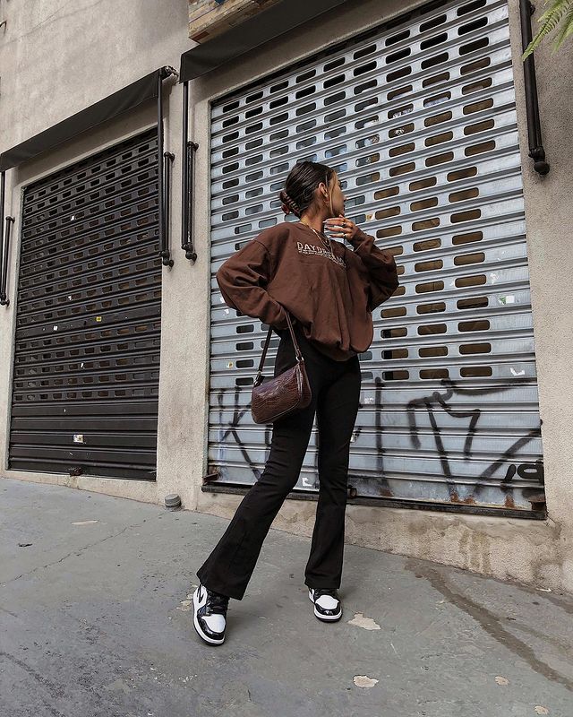 Foto de uma mulher na calçada. Ela veste um moletom vintage marrom, calça bailarina preta e tênis preto e branco. Ela olha para o lado e faz pose.