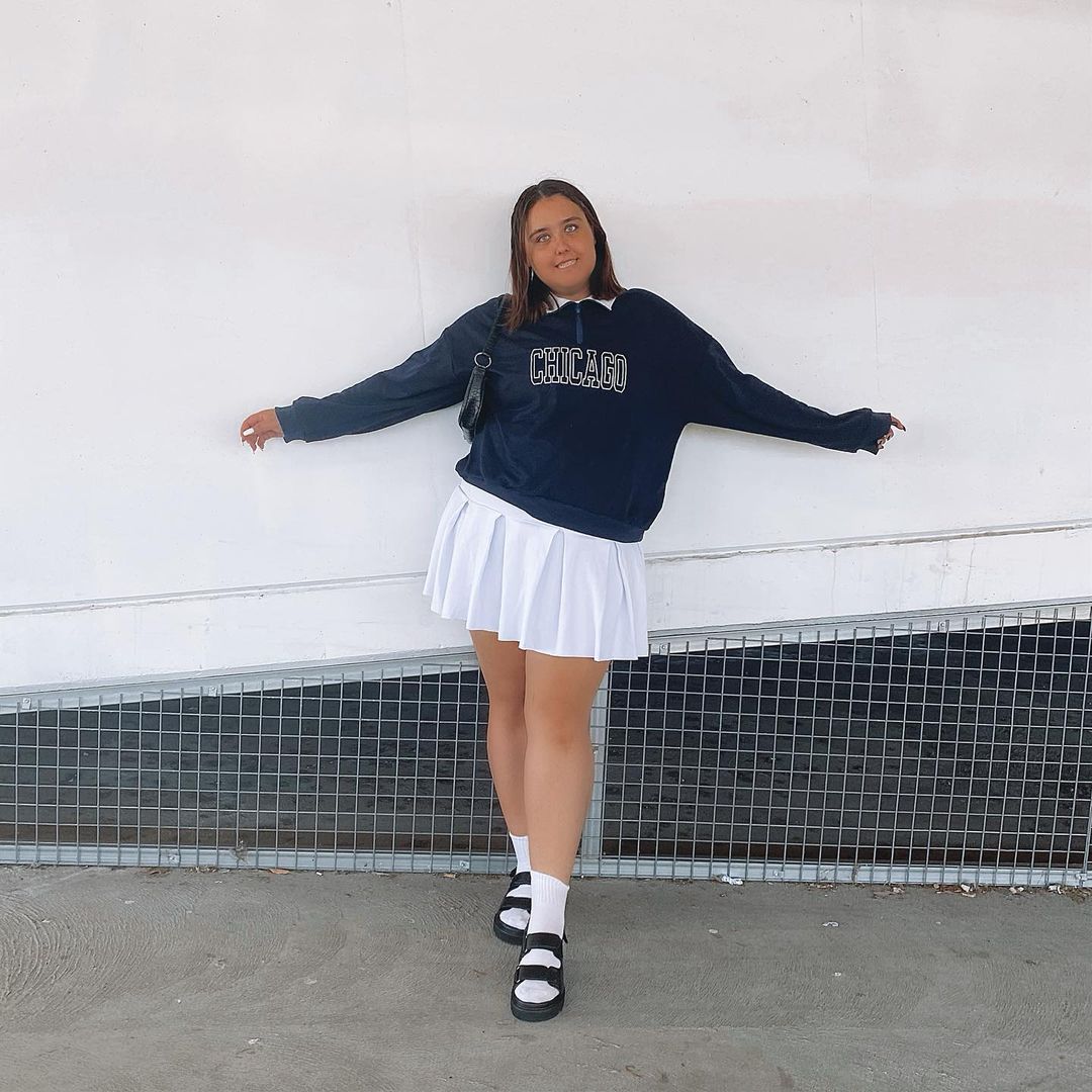 moletom_vintage_saia_colegial Foto de uma mulher com o fundo de uma parede branca. Ela usa um moletom vintage azul escuro, saia colegial branca e papete preta com meia branca. Ela olha para a câmera e sorri.