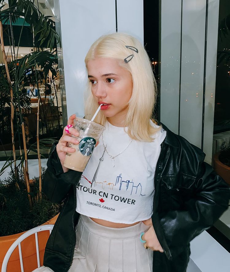 nalim_cabelo_loiro Foto de uma garota. Ela usa um cropped branco, jaqueta de couro preta e saia colegial branca. O cabelo dela está solto, ela segura um copo de café e não olha para a câmera.