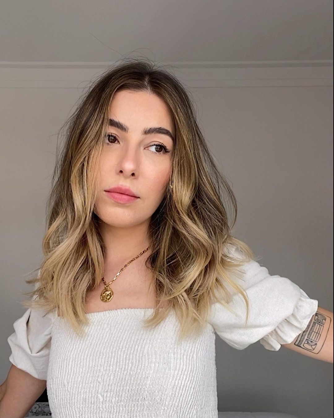 nanaths_cabelo_loiro Selfie de uma mulher. Ela usa uma blusa de manga bufante branca, colar com um medalhão dourado e cabelo loiro solto. Ela olha para o lado e não sorri.
