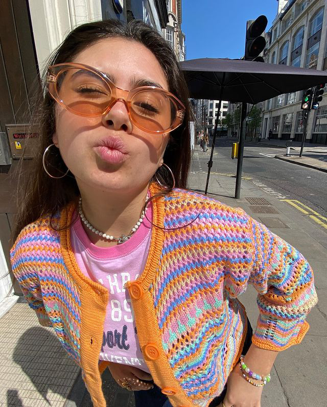 oculos-de-sol-tendencia-lifeasconstance Garota usando camiseta lilás, tricô colorido com laranja, azul e rosa, óculos de sol com armação retrô oval no estilo anos 60. Ela está fazendo um bico com a boca.
