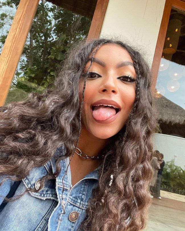 Selfie da Any Gabrielly. Ela está usando uma jaqueta jeans, colar prata, cabelo ondulado solto com um penteado de trança na franja com elástico. Ela olha para a câmera e mostra a língua.