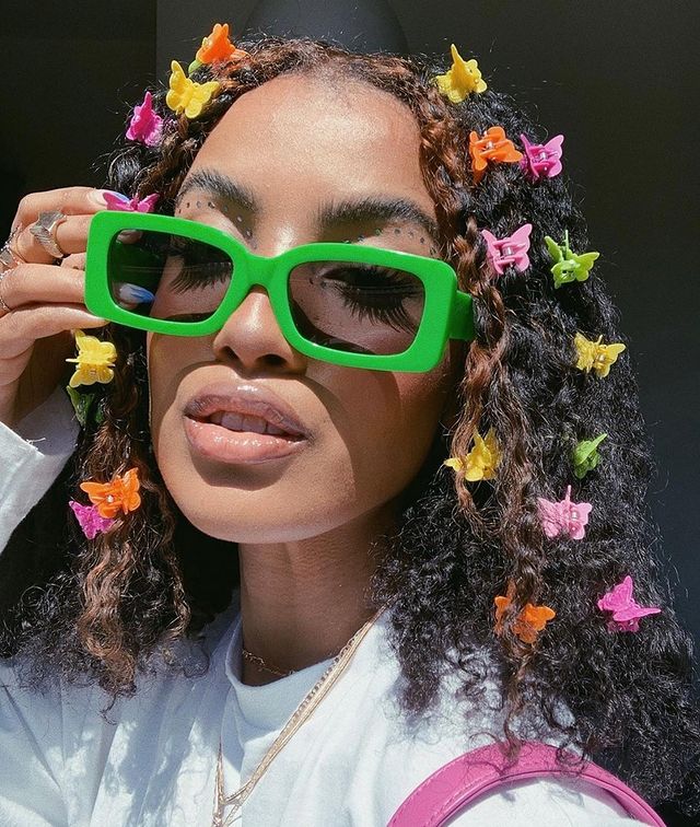 presilha de borboleta laranja rosa e amarela Jovem usando óculos verde com presilha de borboleta colorida no cabelo, com expressão séria.