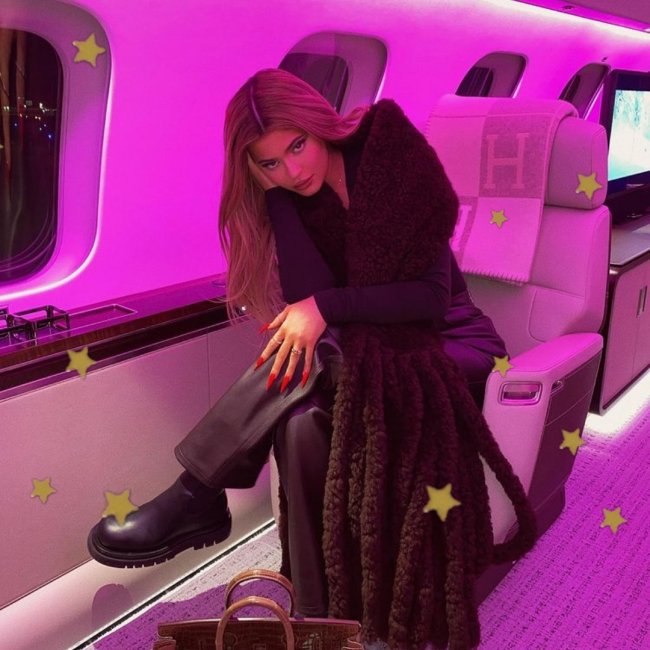 salto tratorado Kylie Jenner posando em avião com bota, uma de suas mãos está apoiando sua cabeça de lado, podemos ver a cadeira do avião e parte da janela.Sua expressão é levemente sorridente.