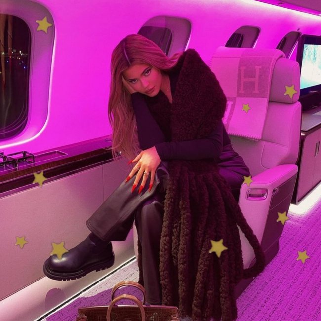 Kylie Jenner posando em avião com bota, uma de suas mãos está apoiando sua cabeça de lado, podemos ver a cadeira do avião e parte da janela.Sua expressão é levemente sorridente.