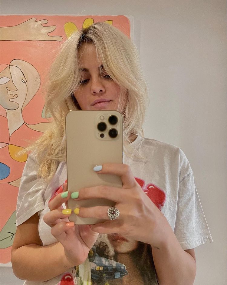 selena_gomez_loira Selfie no espelho da cantora e atriz Selena Gomez. Ela usa uma camiseta branca com estampa vermelha, segura o celular com as duas mãos e está com o cabelo solto. Ela olha para o celular e não sorri para a foto.