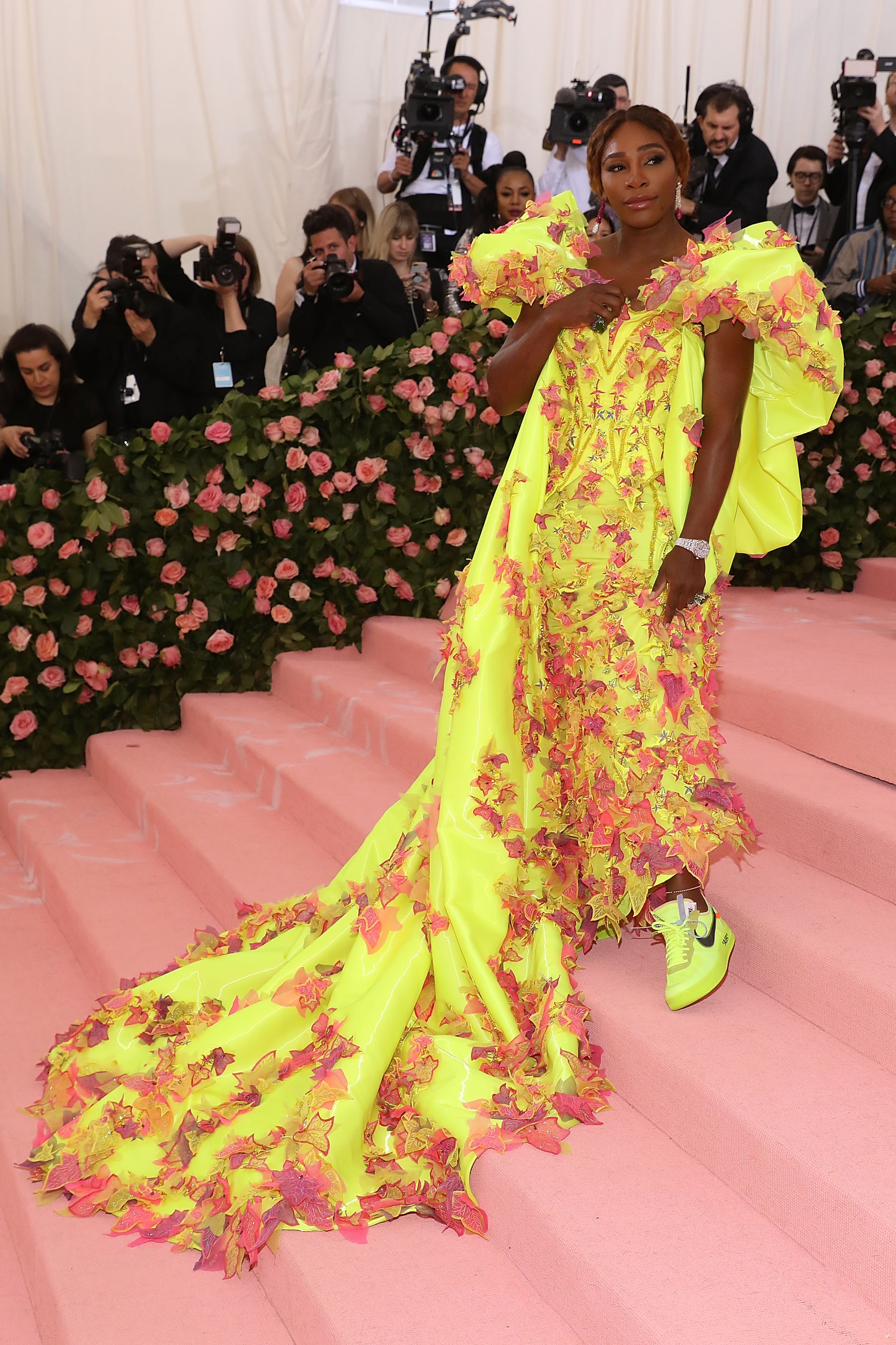 Foto da tenista Serena Williams no tapete vermelho do Met Gala 2019. Ela usa um vestido amarelo longo, de mangas bufantes e detalhes de folhas rosas e tênis amarelo da Nike. Ela olha para o lado e sorri.