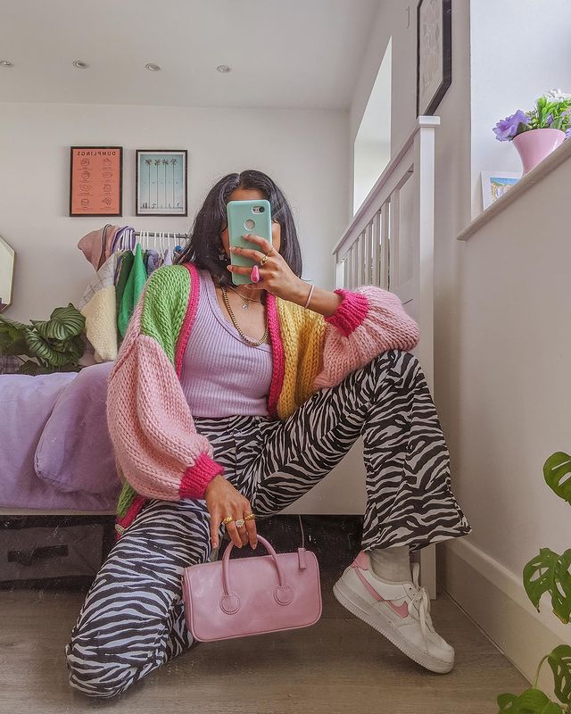 trico-colorido-outono-inverno-poseandrepeat Garota usando look com tricô coloorido em verde, rosa claro, rosa escuro e amarelo, regata lilás, calça preta e branca de zebrinha e tênis branco. Ela está agachada em frente ao espelho, com o celular cobrindo o rosto, uma das mãos segurando uma minibolsa rosa. Um joelho está dobrado e toca o chão, o outro está apoiado em um dos braços.