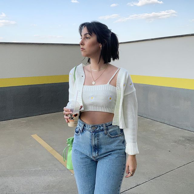 Garota usando top branco com cardigan branco com algumas florzinhas aplicadas e uma calça jeans de cintura alta e lavagem clara. Ela está olhando para o lado, com uma bolsa verde pendurada em um dos ombros e segurando um copo com uma mão.