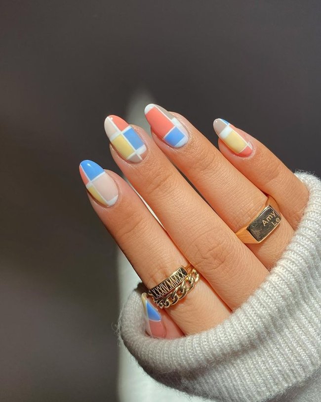unhaxadrezcolorida Foto de uma mão com anéis no dedo anelar e indicador. As unhas estão com uma nail art xadrez nas cores azul, amarela, marrom, nude e salmão.