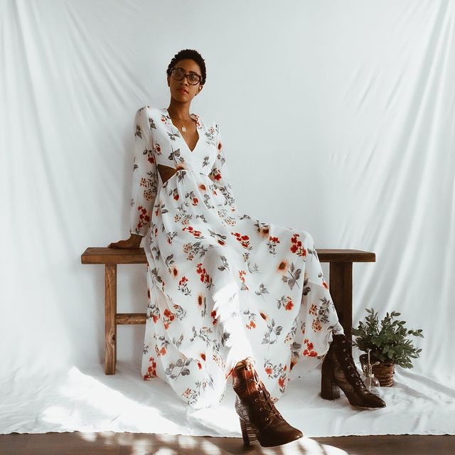 vestido-floral-boho-chic-_soulxflower Garota usando vestido longo floral com fundo branco e botinha marrom com salto e bico fino de verniz. Ela está sentada em um banquinho de madeira, com as duas mãos para trás do corpo. Ao fundo, há um pano branco que serve como cenário para a foto.