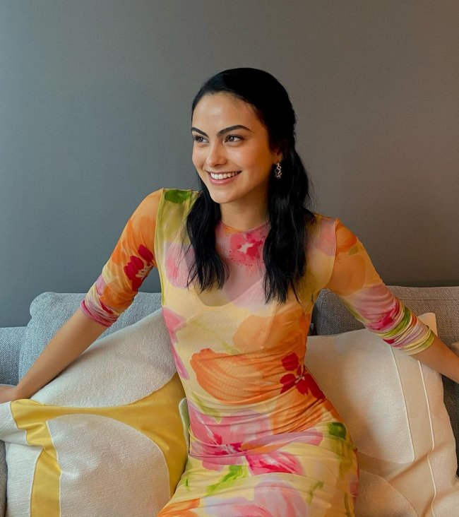 Camila Mendes sentada em sofá branco, com os braços abertos, e expressão sorridente, ela usa vestido semitransparente com estampas nas cores rosa, laranja e verde.