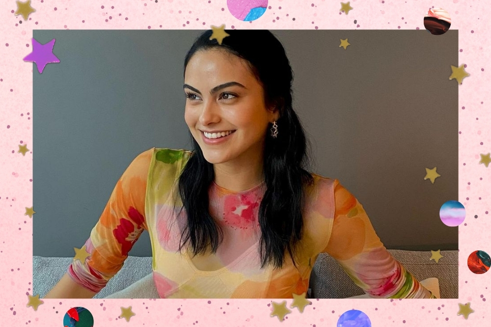 Camila Mendes sentada em sofá branco, com os braços abertos, e expressão sorridente, ela usa vestido semitransparente com estampas nas cores rosa, laranja e verde.