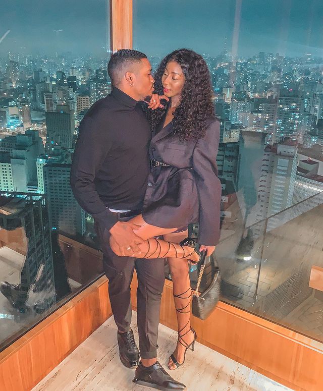Camilla de Lucas e namorado Casal posando em frente a parede de vidro, o rapaz levanta a perna da moça. Os dois estão com uma expressão séria e as roupas que eles vestem são pretas.
