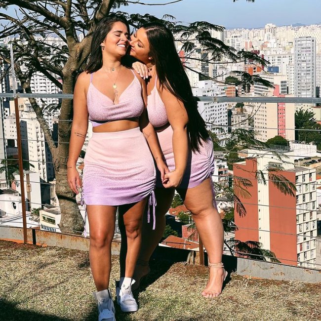 Letticia Muniz e namorada com look combinando Casal posando com uma jovem de frente e a outra de lado a beijando, elas usam a roupa igual. Um top e uma saia rosa claro. Uma usa tênis e a outra uma sandalia de salto. As duas estão com uma expressão alegre.