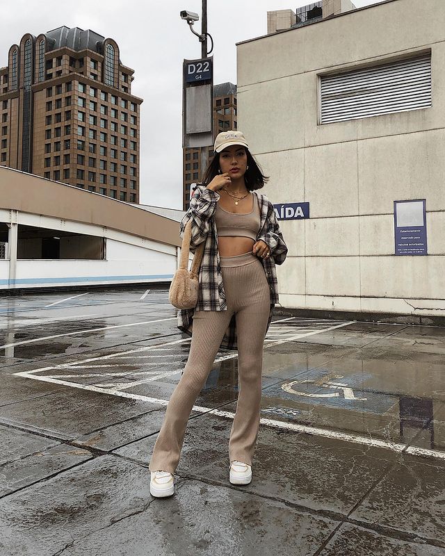 Look calça bailarina e camisa xadrez Foto de uma mulher na rua. Ela usa um top cropped e uma calça legging bailarina bege, camisa xadrez, boné, bolsa bege e tênis branco. Ela olha para a câmera e não sorri.